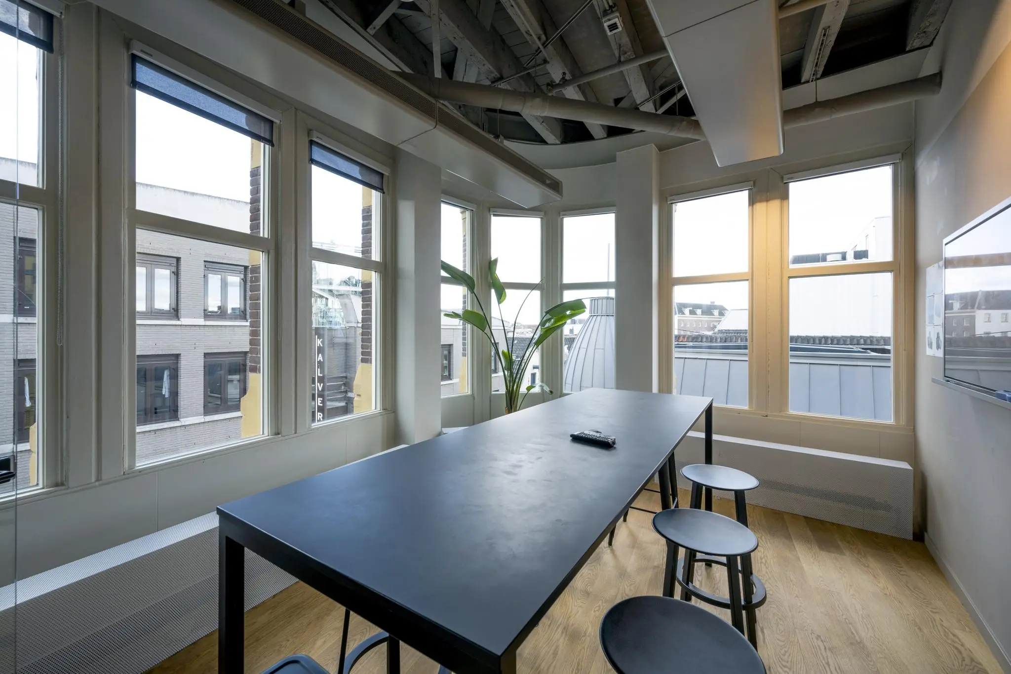 Vergaderruimte met hoge ramen, een zwarte tafel met krukken en uitzicht op gebouwen aan de Sint Jorissteeg in Amsterdam.